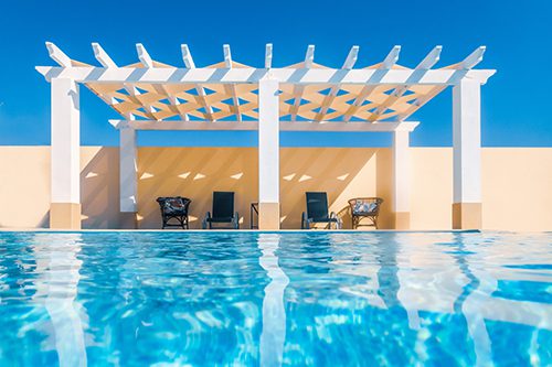 A vinyl pergola provides shade for a seating area next to a pool.